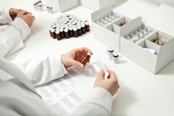 A person in a lab coat holds a small, labeled vial and a document, surrounded by multiple vials and boxes of pharmaceutical supplies on a white table.